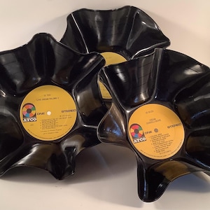 May include: Three black vinyl records shaped into bowls with wavy, flared edges. Each record features a central yellow label with the Atco logo and text including "Cream" and "Life Cream Volume II," displaying track lists and stereo markings.