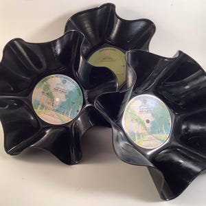May include: Three black, wavy-edged bowls made from repurposed vinyl records. Each bowl features the original record label in the center. The labels have a variety of colors and text, including "Deep Purple" and "Made in Japan."