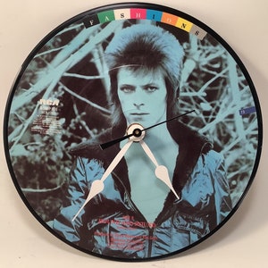 May include: A black, round wall clock featuring a portrait of a person with blue hair and a blue jacket. The clock face is a vintage record album. The word "FASHIONS" is printed at the top of the clock.