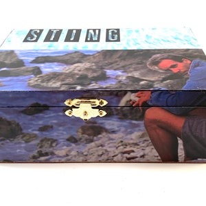 May include: A wooden box with a blue and white image of a man sitting on rocks by the ocean. The box has a gold latch and the word "STING" printed on the top.
