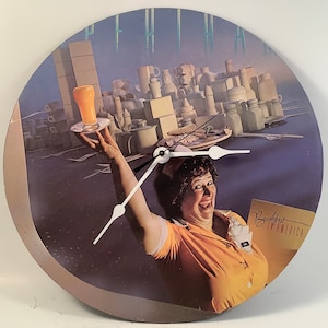 May include: A vintage vinyl record clock featuring a woman in a yellow uniform holding a tray with a glass of orange juice. The clock face is surrounded by a purple background with a white clock hand. The record is titled "Breakfast in America" by Supertramp.