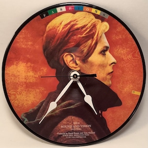 May include: A black vinyl record clock featuring a portrait of David Bowie on the front. The clock face is black with white hands. The record label is for the album "Sound and Vision" by David Bowie. The text "Side A" is printed on the label, along with the album title, artist name, and production information.