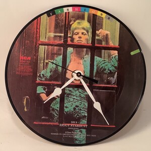 May include: A black vinyl record clock featuring the album cover for David Bowie's "Ziggy Stardust" album. The album cover features a man in a green and black patterned shirt standing behind a window with red and black bars. The clock hands are white and the clock face is black with the album title and artist name printed in white.