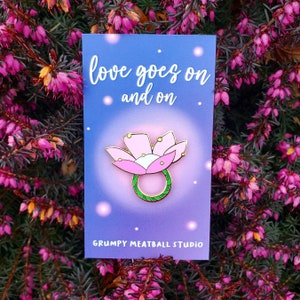 May include: A pink flower with a green ring around it. The flower has five petals and a yellow centre. The background is a blue card with the text "love goes on and on" and "GRUMPY MEATBALL STUDIO".