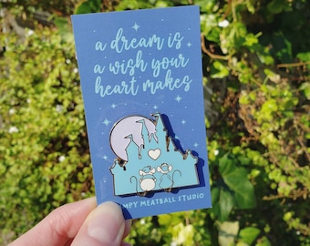Può includere: Uno spillo in smalto blu e bianco con un castello di cartoni animati con due topi davanti. Lo spillo dice "a dream is a wish your heart makes" e "Mpy Meatball Studio".