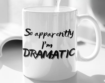 Op de afbeelding: Witte keramische koffiemok met zwarte tekst die "So apparently I'm DRAMATIC" luidt. Een doorzichtige glazen kan giet water in de mok.