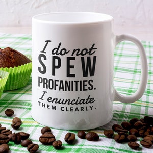 May include: White coffee mug with black text that reads "I do not SPEW PROFANITIES. I enunciate them clearly." The mug is on a green and white checkered tablecloth with chocolate muffins and coffee beans.