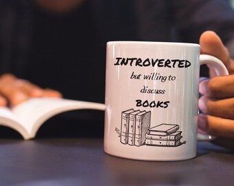 Puede incluir: Taza de cerámica blanca con texto negro que dice "Introvertido pero dispuesto a discutir libros". La taza tiene una ilustración negra de una pila de libros. Una persona está sosteniendo la taza con su mano derecha y leyendo un libro con su mano izquierda.