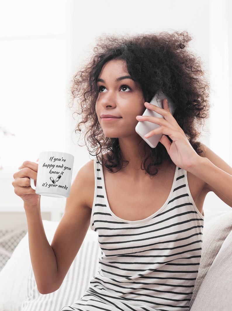 May include: White ceramic mug with black text that reads "If you're happy and you know it it's your meds" with a graphic of two pills. A woman is holding the mug and talking on a white cell phone.