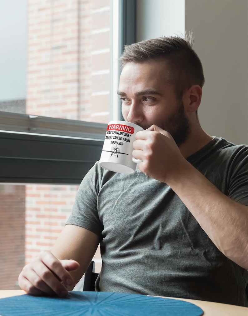May include: White ceramic mug with a red warning sign that reads "WARNING MAY SPONTANEOUSLY START TALKING ABOUT AIRPLANES". A small airplane icon is in the center of the sign. A person is holding the mug and drinking from it.