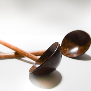 Op de afbeelding: Twee houten pollepels met lange handvatten en diepe, afgeronde kommen. De pollepels zijn donkerbruin van kleur, met een zichtbare natuurlijke houtnerf. De achtergrond is helderwit, wat de vorm en textuur van de pollepels benadrukt.