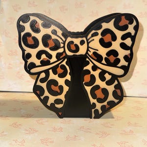 May include: A decorative bow-shaped object with a leopard print design. The bow features a tan background with black spots and brown accents. The edges and the base of the bow are black. The object is likely a decorative item or a holder.