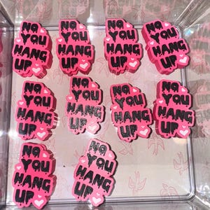 May include: A collection of pink, irregular-shaped stickers with the phrase "No You Hang Up" in black, dripping font. Each sticker has a small white heart at the bottom. The stickers are arranged inside a clear plastic container.