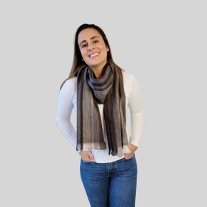 May include: A woman wearing a white long-sleeved shirt and blue jeans is smiling. She is wearing a brown and white striped scarf around her neck.