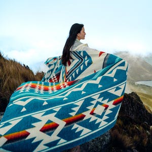 May include: A turquoise, white, and red patterned blanket with geometric designs. The blanket has a colorful stripe of orange, yellow, and red. The blanket is draped over a person outdoors.