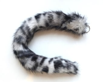 May include: A white and black faux fur tail with a silver ring attached. The tail is curled into a C shape.