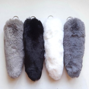 May include: Four plush faux fur keychains arranged in a row. The keychains are coloured light grey, black, white, and dark grey. Each fluffy, rounded accessory features a silver-toned metal split ring attached to the top for easy hanging or attachment.
