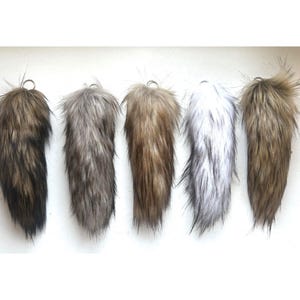 May include: Five faux fur tails arranged in a row, each featuring a metal ring at the top. The colours range from dark brown and grey to light tan, white with black tips, and a golden brown, all displaying long, textured synthetic fibres.