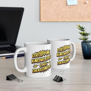 May include: Two white ceramic mugs with the text "DROPPING KNOWLEDGE LIKE DRAGONFIRE" in yellow and black. The mugs are on a light-colored surface, with a laptop and office supplies in the background.