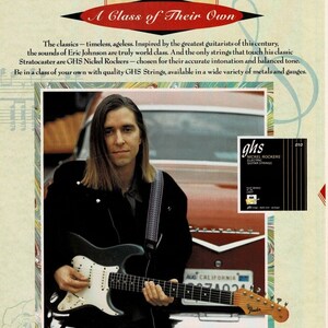 May include: A musician plays a blue electric guitar with a white pickguard. The guitar has a silver Fender logo on the headstock. The musician is wearing a black jacket and a striped guitar strap. The image is a product advertisement for GHS Nickel Rockers electric guitar strings.