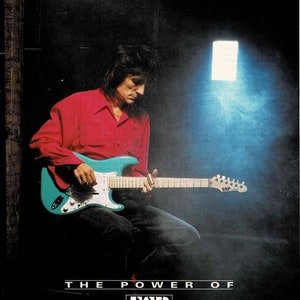 May include: A musician plays a teal electric guitar with a white pickguard. The guitar has a black ESP logo on the headstock. The musician is wearing a red shirt and black pants. The text "THE POWER OF ESP" is printed below the guitar. The text "Ronnie Wood plays the ESP Traditional Series on his latest release, "Slide on This" from Continuum Records." is printed below the text "THE POWER OF ESP".