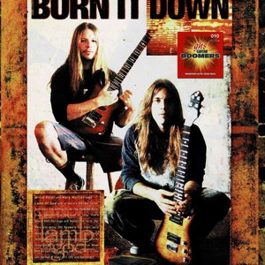 May include: Two musicians, one with long blonde hair and one with long brown hair, are playing electric guitars. The musicians are wearing casual clothing and are standing in front of a red, white, and blue background. The text "BURN IT DOWN" is written in large, bold letters at the top of the image. The text "GHS Strings and ROCKTRON featured artist - LAMB OF GOD" is written in smaller letters below the main text. The image is a promotional advertisement for GHS Strings and ROCKTRON.