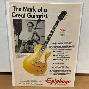 May include: A gold Les Paul Standard electric guitar with a gold top. The guitar is made by Epiphone and features a rosewood fingerboard with trapezoid inlays. The ad text reads "The Mark of a Great Guitarist." and "New! Les Paul Standard Gold Top".