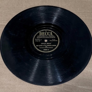 May include: A black circular vinyl record featuring a Decca Personality Series label in the center. The label displays the text Jingle Bells, Bing Crosby and the Andrews Sisters with Vic Schoen and His Orchestra, and the catalog number 23281 A.