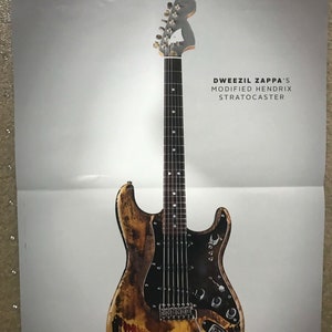 May include: A close-up of a worn, yellowed, and red-stained electric guitar with a black pickguard. The guitar has a black neck with silver tuning pegs and a white pickguard. The text "Dweezil Zappa's Modified Hendrix Stratocaster" is printed in black on a white background above the guitar. The text "Guitar World Collector's Choice" is printed in black on a white background below the guitar.