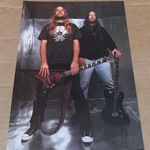 May include: Two men with long hair are standing in a room with a metal wall. They are both holding electric guitars. The man on the left is wearing a black t-shirt with the text "RPG" on it. The man on the right is wearing a black shirt and blue jeans.