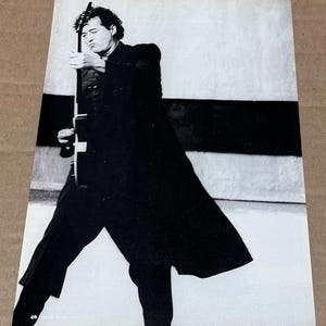 May include: Black and white photograph of a person holding a guitar. The person is wearing a long black coat and pants. The background has a white and black stripe design. The image is a portrait.