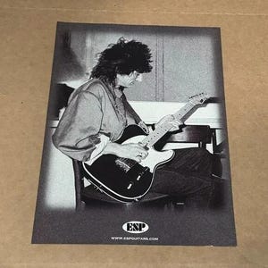 May include: Black and white photograph of a person playing a black electric guitar with a white pickguard. The guitar has an "ESP" logo on the headstock. The text "www.espguitars.com" is printed below the logo.