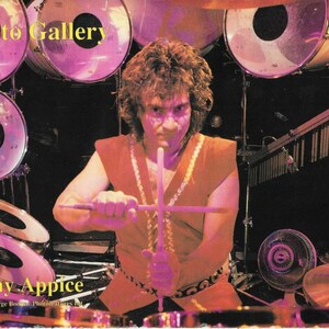Puede incluir: Un baterista, Vinny Appice, está tocando una batería con baquetas. Los tambores son de color rosa y tienen bordes negros. El baterista lleva una camisa roja con ribetes dorados y un cinturón negro. La foto está subtitulada con "Photo Gallery" y "Vinny Appice".