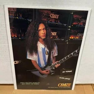 May include: A musician is playing a black electric guitar with a white pickguard. The guitar has a Crate logo on the headstock. The musician is wearing a blue and white striped shirt and is standing in front of a wall of amplifiers. The text "On Stage With Crate" is visible in the lower right corner of the image.