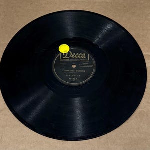 Op de afbeelding: Zwarte vinylplaat met een geel label. Het label heeft de tekst "Decca" en "Tennessee Border" door "Red Foley" met het nummer "46181-A".