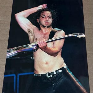 May include: A live performance photograph of a musician playing an electric guitar. The musician is shirtless, wearing black leather pants and a belt with a silver buckle. The guitar has a metallic finish.