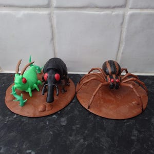 May include: Two circular brown bases featuring insect figurines. The left base holds a bright green grasshopper-like creature and a black beetle, both with red eyes. The right base features a brown spider with black stripes and red eyes.