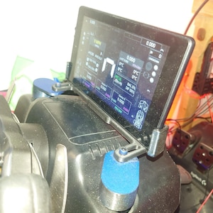 May include: A black digital dashboard display mounted on a racing steering wheel base. The screen shows various telemetry data including speed in MPH, gear indicator, and sector times. The unit is secured with two blue cylindrical mounts.