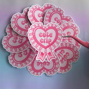 Puede incluir: Un montón de pegatinas en forma de corazón rosa con el texto "cute crip" en una fuente retro. Las pegatinas tienen un borde blanco y un volante rosa alrededor del borde.