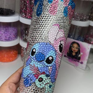 Puede incluir: Un vaso adornado con pedrer&iacute;a con los personajes Stitch y Angel de Lilo & Stitch. El vaso est&aacute; cubierto de pedrer&iacute;a plateada con detalles azules y rosas. Los personajes est&aacute;n representados con pedrer&iacute;a de colores.