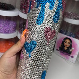 Puede incluir: Un vaso adornado con pedrer&iacute;a con una base plateada, adornado con dise&ntilde;os de corazones y gotas azules y rosas. El vaso est&aacute; cubierto de peque&ntilde;os y brillantes pedrer&iacute;a. El fondo incluye recipientes de pedrer&iacute;a de varios colores.