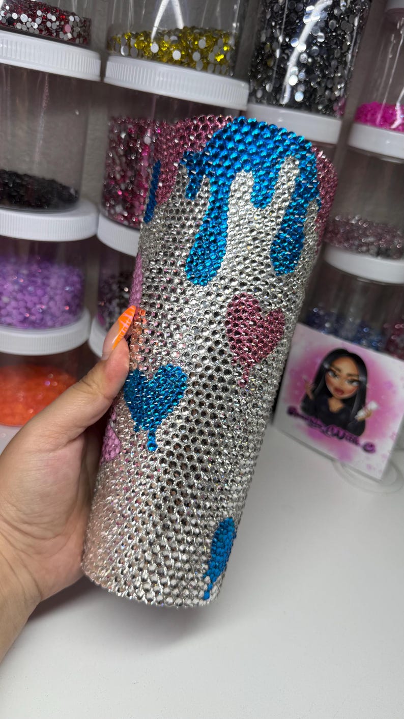 Puede incluir: Un vaso adornado con pedrer&iacute;a con una base plateada, adornado con dise&ntilde;os de corazones y gotas azules y rosas. El vaso est&aacute; cubierto de peque&ntilde;os y brillantes pedrer&iacute;a. El fondo incluye recipientes de pedrer&iacute;a de varios colores.