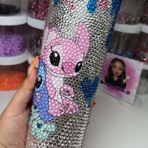 Puede incluir: Un vaso plateado cubierto de pedrer&iacute;a, con un dise&ntilde;o de personaje de dibujos animados rosa y azul. La parte superior del vaso tiene un efecto de goteo con pedrer&iacute;a rosa y azul. El vaso es cil&iacute;ndrico.