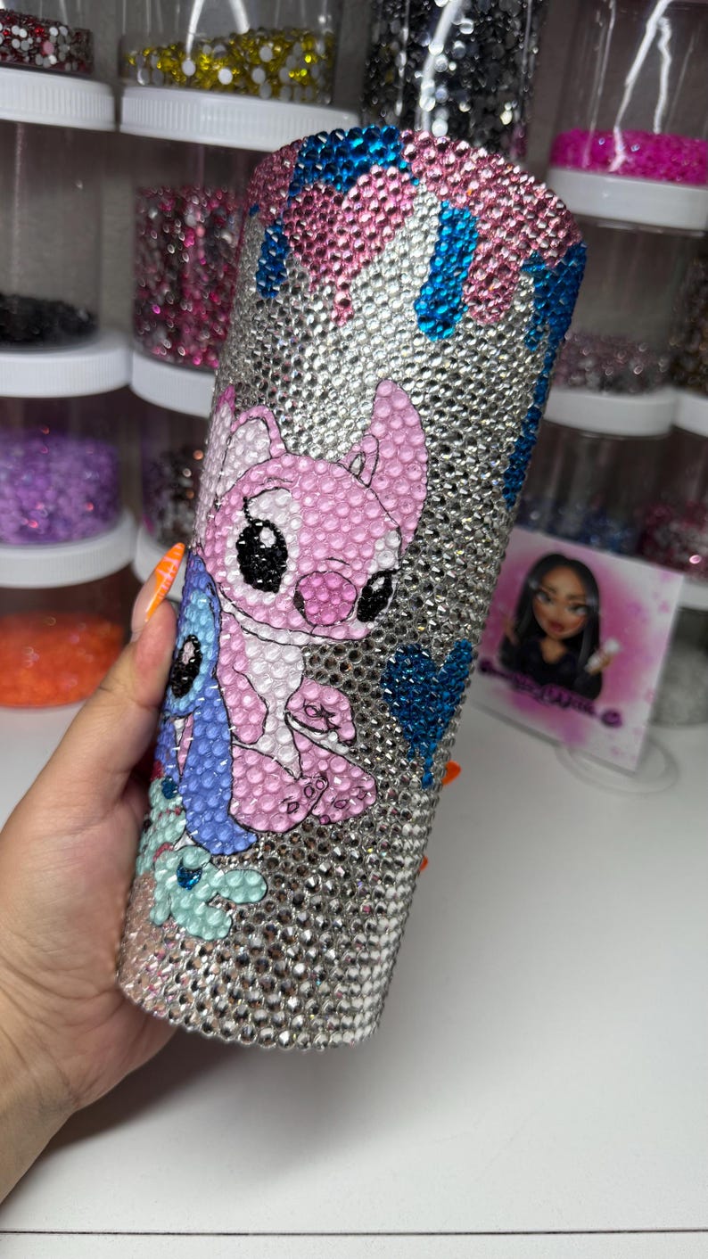 Puede incluir: Un vaso plateado cubierto de pedrer&iacute;a, con un dise&ntilde;o de personaje de dibujos animados rosa y azul. La parte superior del vaso tiene un efecto de goteo con pedrer&iacute;a rosa y azul. El vaso es cil&iacute;ndrico.