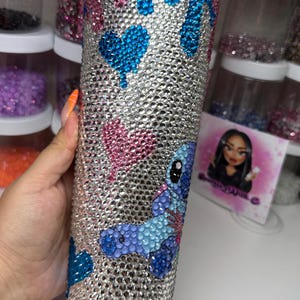 Puede incluir: Un vaso adornado con pedrer&iacute;a con una base plateada, adornado con corazones azules, corazones rosas y un dise&ntilde;o de personaje de dibujos animados azul y morado. El vaso est&aacute; cubierto de brillantes pedrer&iacute;a, creando un efecto brillante. El dise&ntilde;o probablemente est&eacute; inspirado en un personaje de dibujos animados popular.