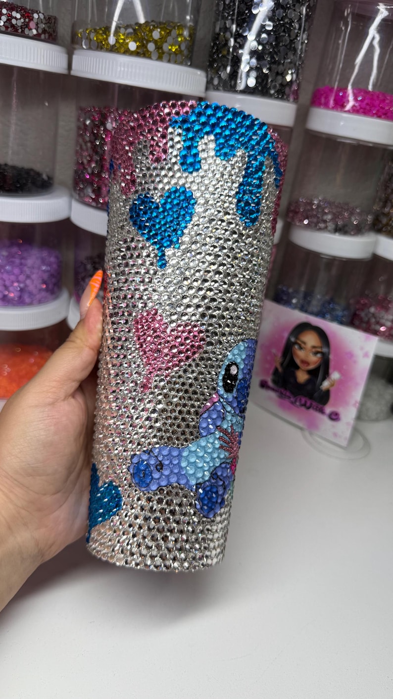 Puede incluir: Un vaso adornado con pedrer&iacute;a con una base plateada, adornado con corazones azules, corazones rosas y un dise&ntilde;o de personaje de dibujos animados azul y morado. El vaso est&aacute; cubierto de brillantes pedrer&iacute;a, creando un efecto brillante. El dise&ntilde;o probablemente est&eacute; inspirado en un personaje de dibujos animados popular.