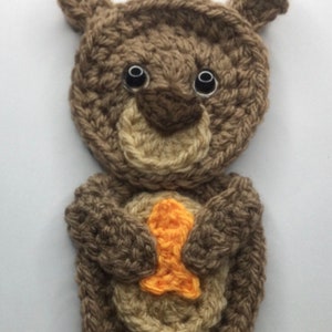 May include: A crocheted otter applique in shades of brown, tan, and orange. The otter has a round body, small ears, and holds an orange fish. The eyes are black. This handmade item is suitable for various craft projects.