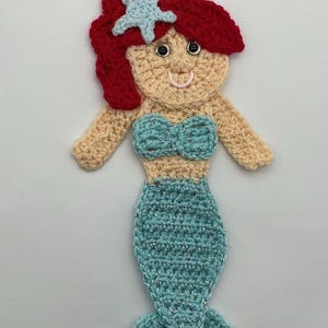 May include: Handmade crocheted mermaid applique. The mermaid has red hair with a light blue starfish, a blue top, and a blue tail. The colours are red, light blue, and beige. A charming craft item.