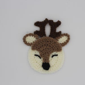 May include: A crocheted deer head with a cream-colored face, brown antlers, and brown ears. The deer has closed black eyes and a black nose. The handmade item is likely a decorative applique or embellishment.