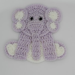 May include: A handmade crochet elephant in lavender and white. The elephant has large, rounded ears with white accents, a long trunk, and small, black googly eyes. The elephant is sitting with its legs spread.